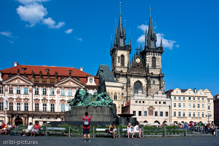 *Prag - Jan-Hus-Denkmal*<br><br>Anfang des 20. Jahrhunderts konzipiert und 1915 anläßlich des 500. Todestages des bedeutenden tschechischen Reformators Jan Hus (1369-1415) fertiggestellt, zählt das mächtige Denkmal zu den wichtigsten Jugendstil-Plastiken Prags. Heute in erster Linie Treffpunkt von Touristen, die sich auf seinen Stufen niederlassen, um das quirlige Treiben auf dem Platz zu verfolgen, war es geschaffen worden als Symbol der nationalen Eigenständigkeit des tschechischen Volkes. Beinahe übermenschlich ragt die Gestalt von Hus aus den übrigen Personen heraus, jener Reformator, der die katholische Kirche scharf kritisiert hatte und deshalb 1415 beim Konstanzer Konzil auf dem Scheiterhaufen verbrannt wurde. Bildhauer des Denkmals ist Ladislav Saloun, dessen Werk häufig mit Plastiken Rodins verglichen wird.<br><br>